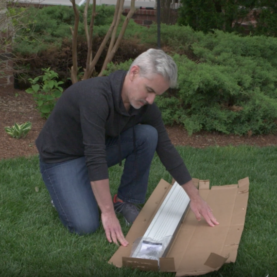 A man with gray hair kneels on grass, opening a large cardboard box containing a long white object—possibly featuring stainless steel micro-mesh—in a garden surrounded by shrubs, plants, and scattered pine needles.